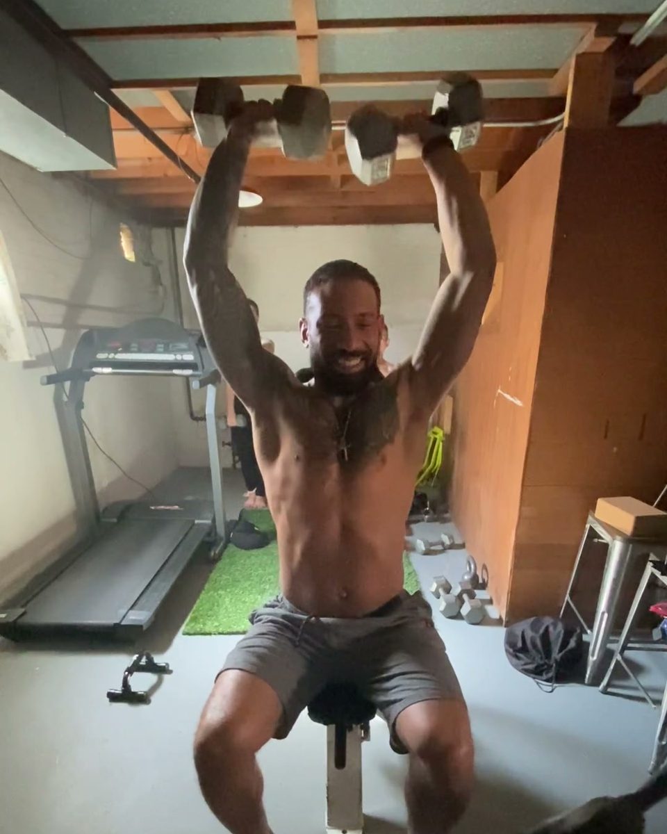 Eddie pressing two dumbbells overhead from a seated position in his home gym